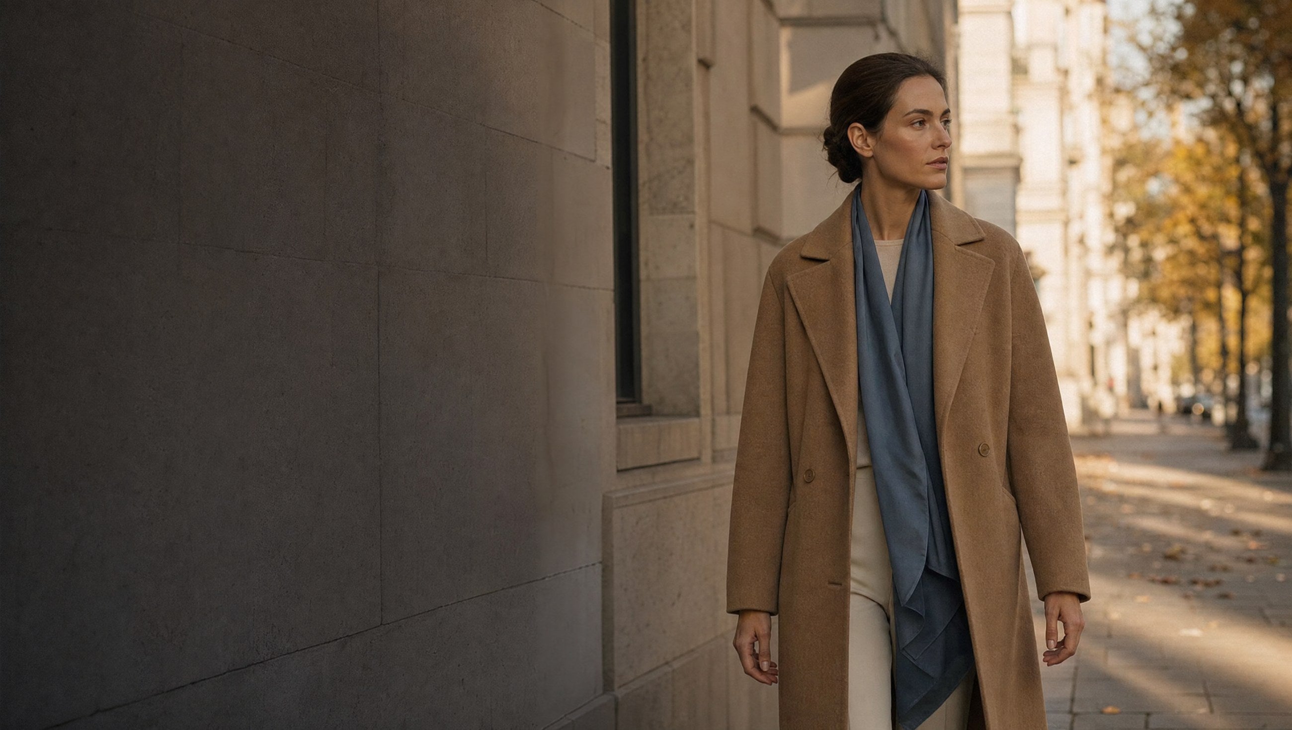 A woman in a camel overcoat walking a limestone street in late autumn light